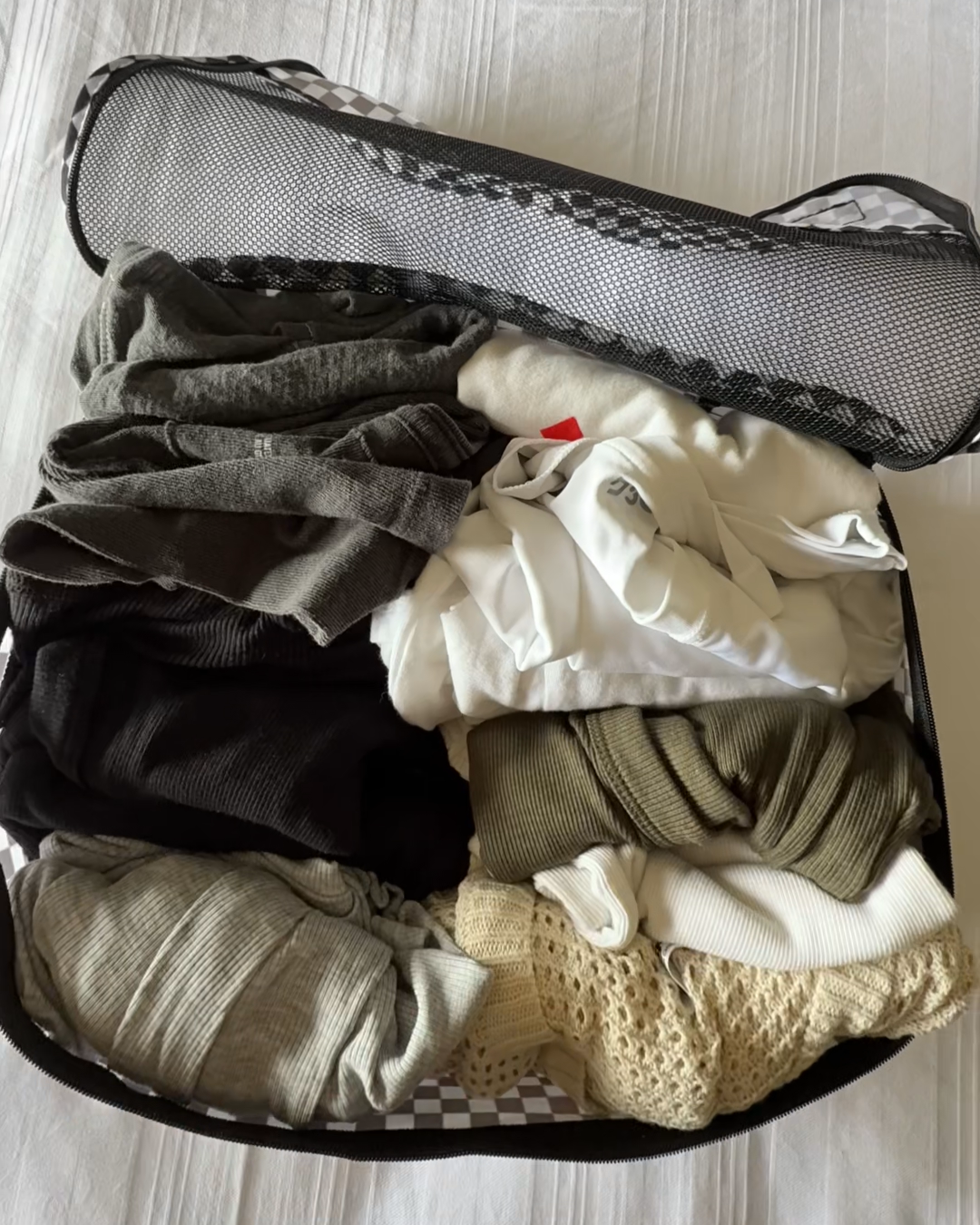 Checkered black and white packing cube filled with folded, rolled clothes on a bed.